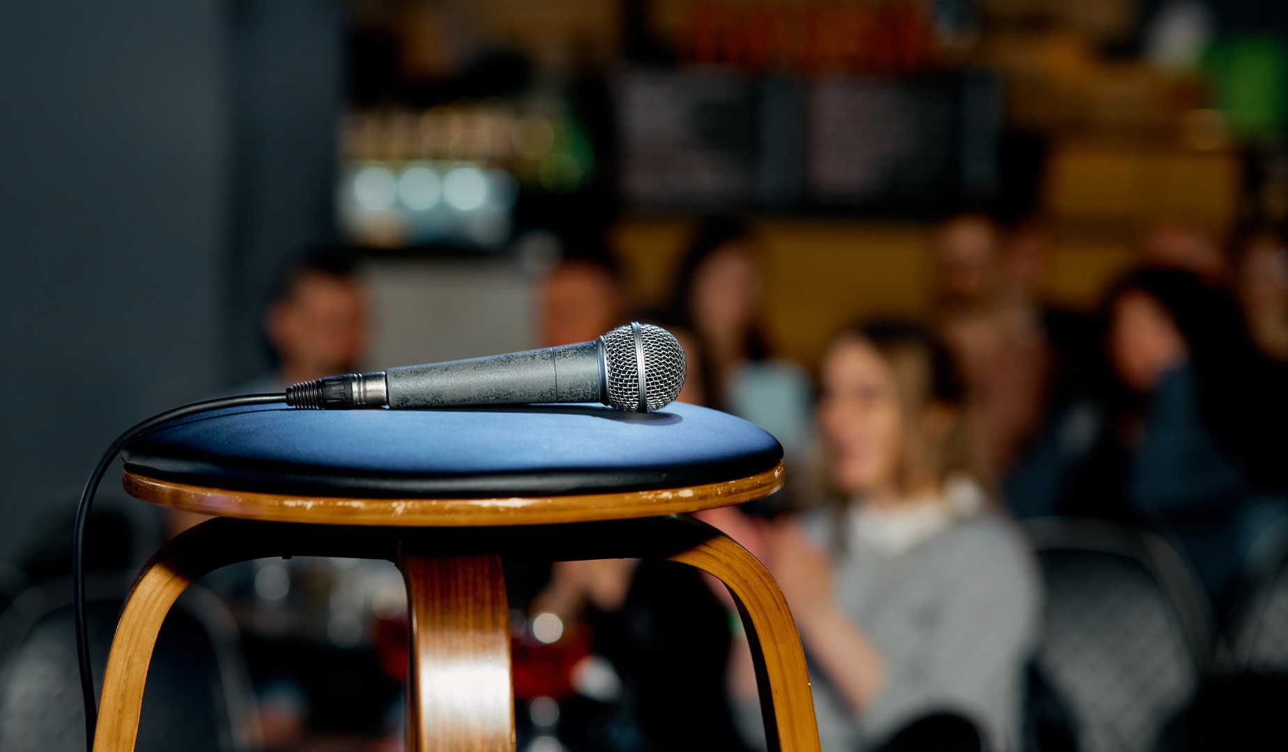 A microphone lies on a spotlit stool as a crowd gathers for a stand-up show at a local comedy club. 